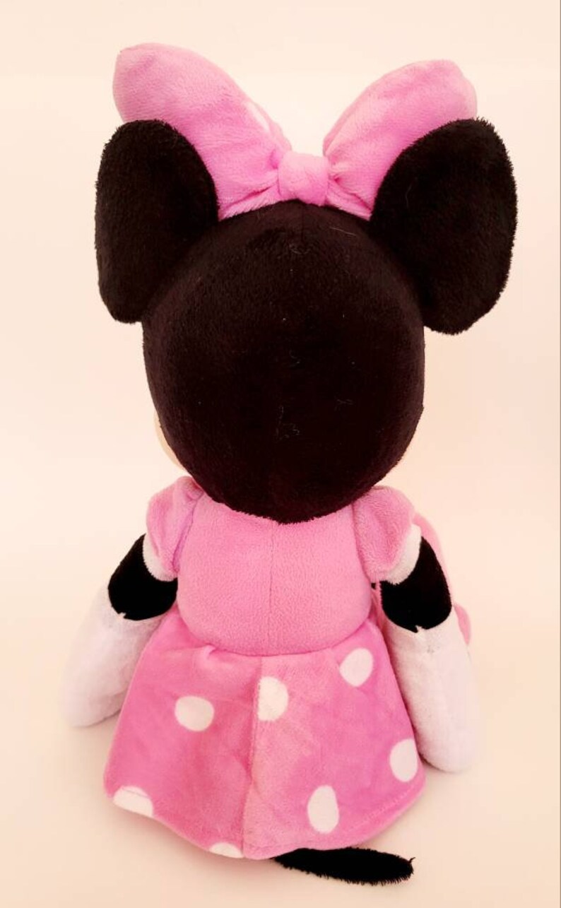 Op de afbeelding: Een pluche Minnie Mouse pop, van achteren gezien, met een zwarte kop, grote oren en een roze strik. De pop draagt een roze jurk met witte stippen en heeft zwart-witte accenten op de armen.