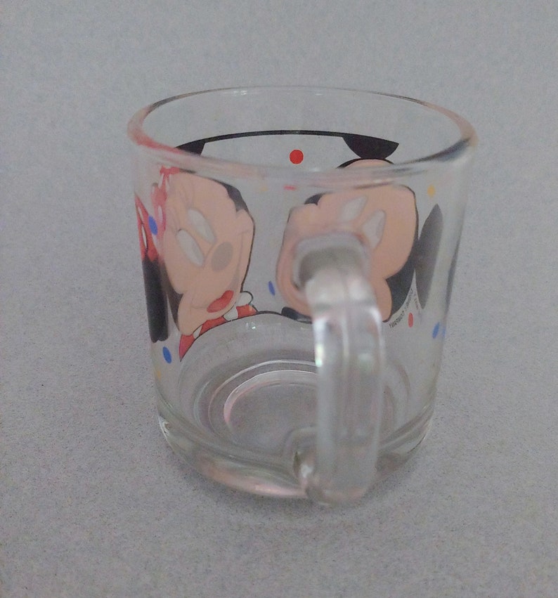 May include: A clear glass mug with a handle. The mug has a cartoon image of Mickey Mouse and Minnie Mouse on it. The image is surrounded by colorful polka dots.