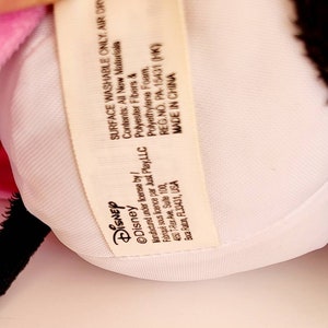 Op de afbeelding: Een close-up van een pluche speelgoed met een witte binnenkant en een roze en zwarte buitenkant. Een label met het Disney-logo en tekst is zichtbaar. Op het label staat "SURFACE WASHABLE ONLY, AIR DRY".