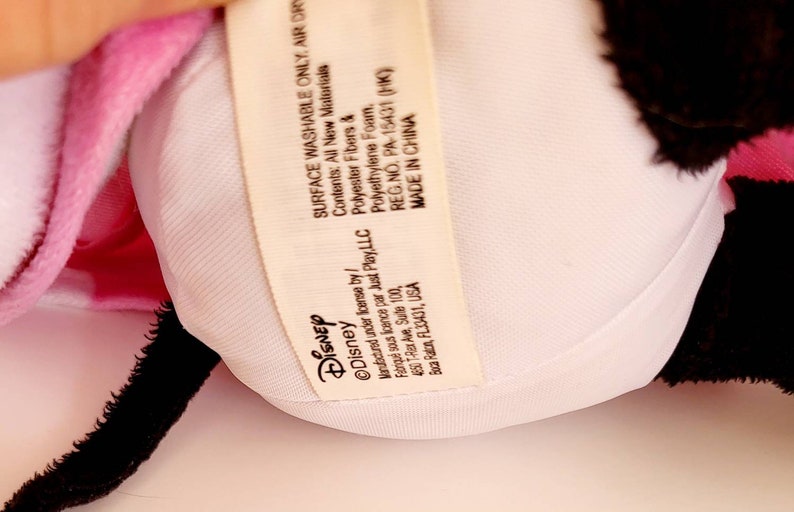 Op de afbeelding: Een close-up van een pluche speelgoed met een witte binnenkant en een roze en zwarte buitenkant. Een label met het Disney-logo en tekst is zichtbaar. Op het label staat "SURFACE WASHABLE ONLY, AIR DRY".