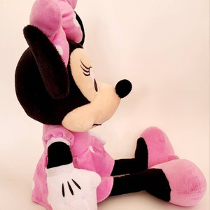 Op de afbeelding: Een zachte Minnie Mouse pop in zittende positie. De pop heeft een zwart hoofd en oren, een roze strik met witte stippen en een roze jurk. Witte handschoenen en roze schoenen zijn zichtbaar.