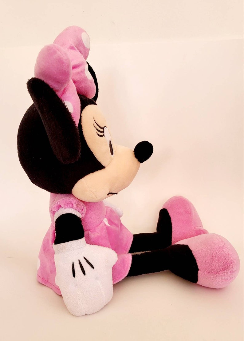 Op de afbeelding: Een zachte Minnie Mouse pop in zittende positie. De pop heeft een zwart hoofd en oren, een roze strik met witte stippen en een roze jurk. Witte handschoenen en roze schoenen zijn zichtbaar.