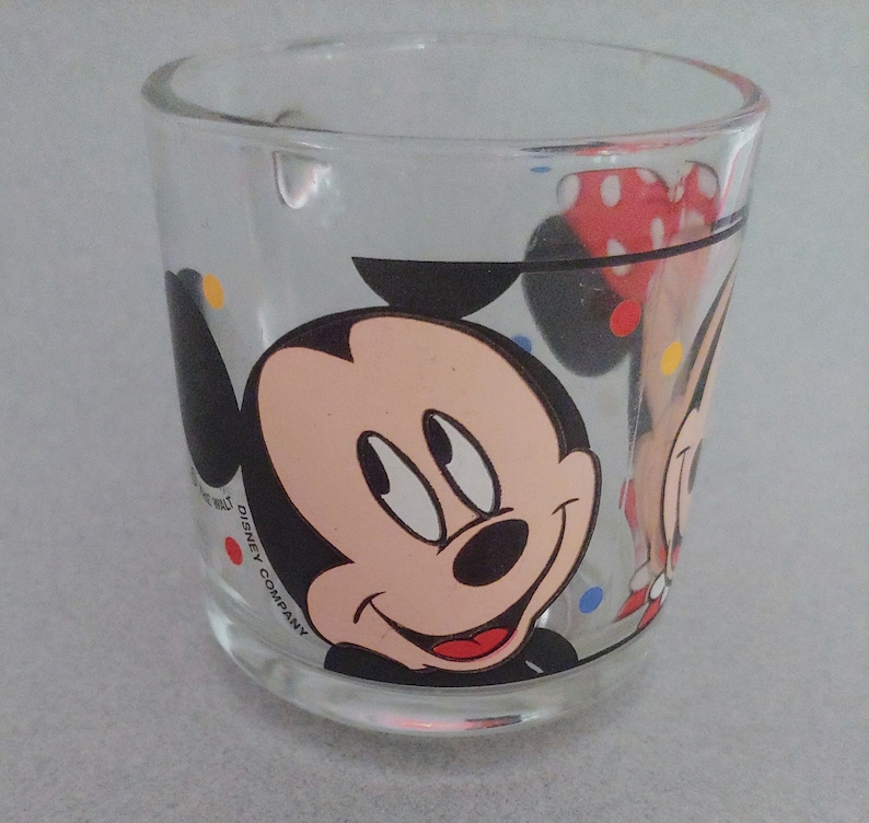 May include: A clear glass mug with a colorful polka dot pattern and a cartoon image of Mickey Mouse and Minnie Mouse. The mug has the text "DISNEY COMPANY" printed on the side.