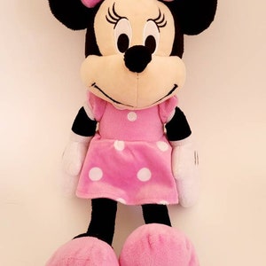 Op de afbeelding: Een Minnie Mouse knuffelpop, gekleed in een roze jurk en strik met witte stippen. De pop heeft zwarte oren, een zwarte neus en witte handschoenen. Grote roze voeten maken het ontwerp compleet.