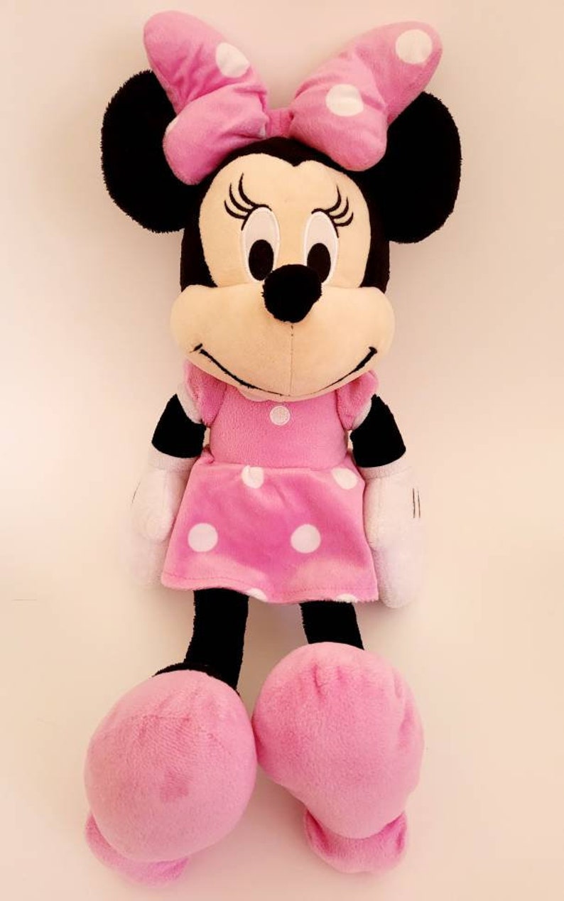 Op de afbeelding: Een Minnie Mouse knuffelpop, gekleed in een roze jurk en strik met witte stippen. De pop heeft zwarte oren, een zwarte neus en witte handschoenen. Grote roze voeten maken het ontwerp compleet.