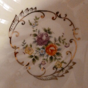 May include: A white ceramic bowl with a gold floral design. The design features pink, yellow, and white flowers with green leaves.