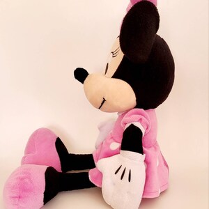Op de afbeelding: Een zittende Minnie Mouse pluche pop. De pop heeft een zwarte kop met grote oren, een roze strik en een roze jurk met witte stippen. Haar handschoenen zijn wit en haar schoenen zijn roze en zwart.
