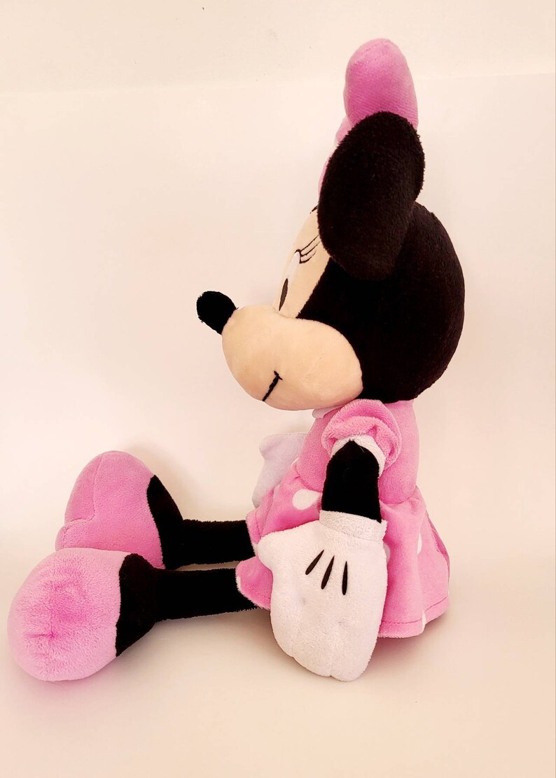 Op de afbeelding: Een zittende Minnie Mouse pluche pop. De pop heeft een zwarte kop met grote oren, een roze strik en een roze jurk met witte stippen. Haar handschoenen zijn wit en haar schoenen zijn roze en zwart.