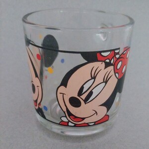 May include: A clear glass mug with a black outline and red polka dots. The mug features a cartoon image of Minnie Mouse with a red bow and a black nose. The background is filled with colorful confetti.