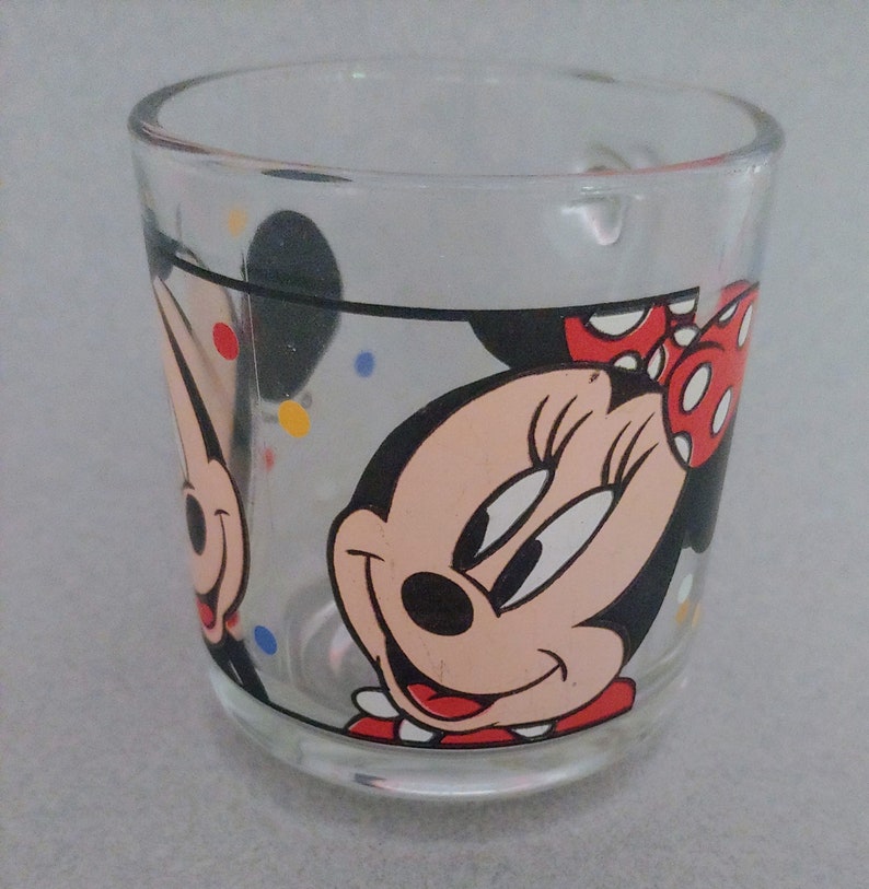 May include: A clear glass mug with a black outline and red polka dots. The mug features a cartoon image of Minnie Mouse with a red bow and a black nose. The background is filled with colorful confetti.