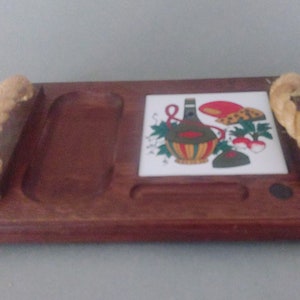 May include: A wooden serving tray with rope handles. The tray has a small rectangular compartment and a larger oval compartment. A white ceramic tile with a colorful illustration of a bottle of wine, mushrooms, and vegetables is attached to the tray.
