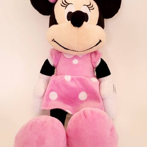 Op de afbeelding: Een Minnie Mouse knuffel. De pop heeft zwarte oren, een roze strik met witte stippen en een roze jurk met witte stippen. De armen en benen van de pop zijn roze en ze heeft een witte kraag.