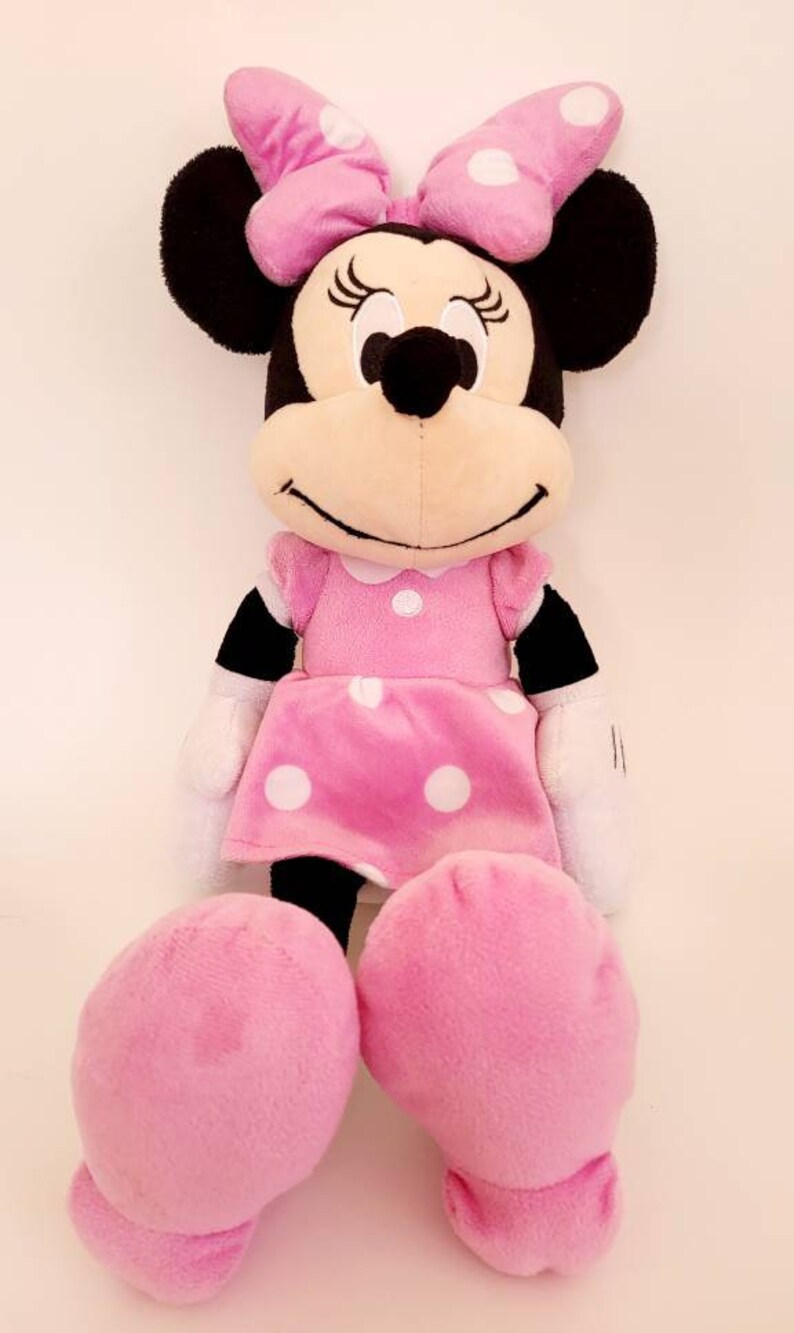 Op de afbeelding: Een Minnie Mouse knuffel. De pop heeft zwarte oren, een roze strik met witte stippen en een roze jurk met witte stippen. De armen en benen van de pop zijn roze en ze heeft een witte kraag.