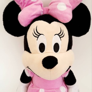 Op de afbeelding: Een pluche Minnie Mouse pop, met een roze jurk en strik met witte stippen. De pop heeft zwarte oren, een zwarte neus en geborduurde wimpers. Dit knuffel is een populair karakter voor kinderen.