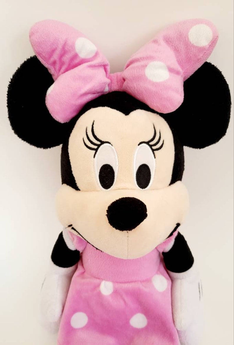 Op de afbeelding: Een pluche Minnie Mouse pop, met een roze jurk en strik met witte stippen. De pop heeft zwarte oren, een zwarte neus en geborduurde wimpers. Dit knuffel is een populair karakter voor kinderen.