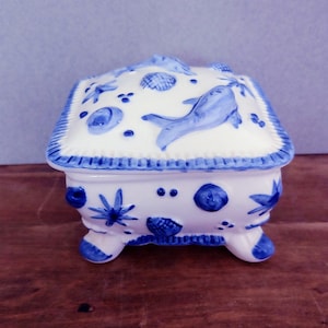May include: A small, white ceramic trinket box with a blue and white design. The box features a raised design of fish and seashells. The box has four legs and a hinged lid.