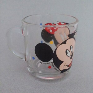 May include: A clear glass mug with a colorful Mickey Mouse and Minnie Mouse design. The mug has a handle and features the text "&copy; THE WALT DISNEY COMPANY".