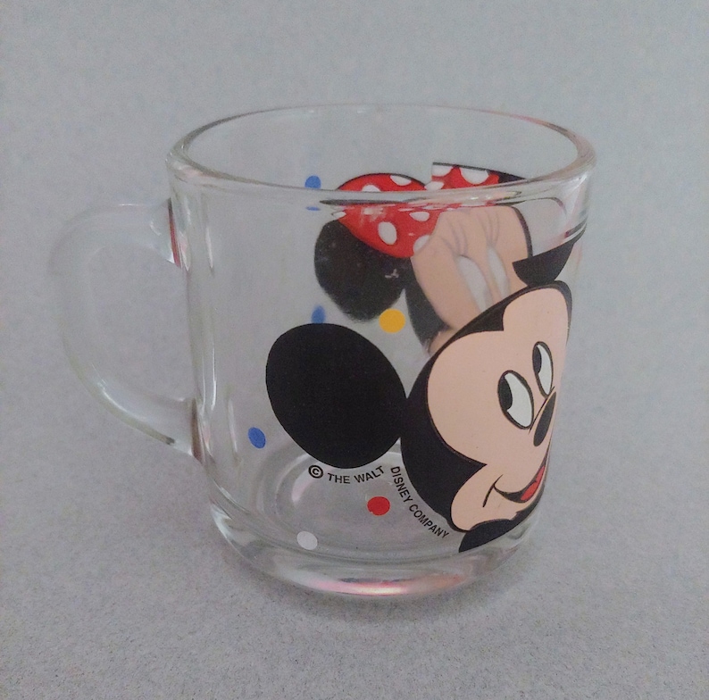 May include: A clear glass mug with a colorful Mickey Mouse and Minnie Mouse design. The mug has a handle and features the text "&copy; THE WALT DISNEY COMPANY".