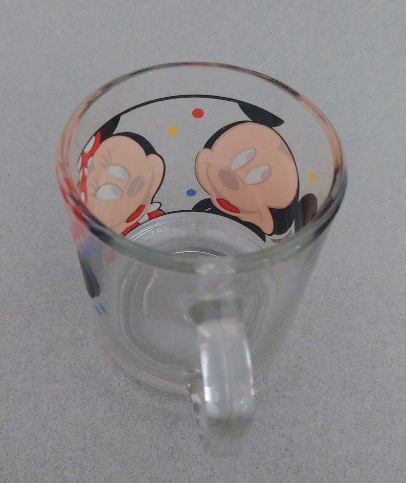 May include: A clear glass mug with a handle. The mug has a colorful design featuring Mickey Mouse and Minnie Mouse.