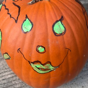 May include: A carved orange pumpkin with a green painted face. The face has two teardrop-shaped eyes, a small nose, and a wide smile.