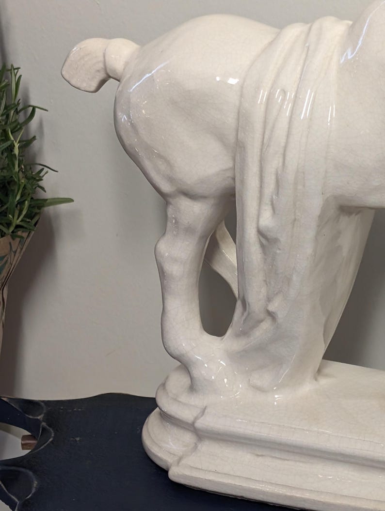 Puede incluir: Estatua de caballo de cer&aacute;mica blanca, que muestra las patas traseras y la tela drapeada. La escultura tiene un acabado brillante y se asienta sobre una base escalonada. La cola del caballo es visible.