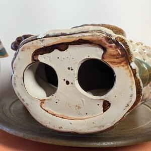 May include: A ceramic owl figurine, viewed from the bottom, showcasing two large circular openings. The owl is primarily beige and brown, with accents of green and white. The base is a speckled off-white color, and the figurine rests on a brown plate.