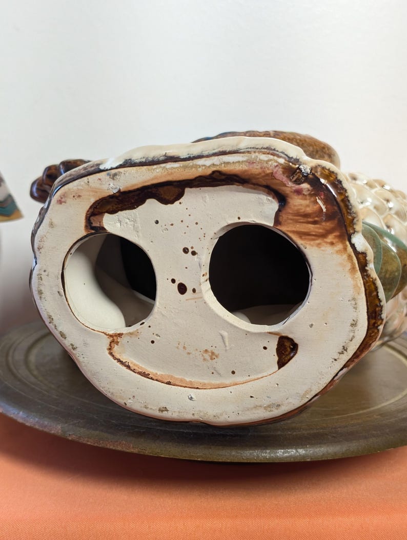 May include: A ceramic owl figurine, viewed from the bottom, showcasing two large circular openings. The owl is primarily beige and brown, with accents of green and white. The base is a speckled off-white color, and the figurine rests on a brown plate.