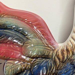 May include: Close-up of a ceramic rooster with a glossy finish. The rooster features a vibrant color palette, including red, blue, green, and brown. The details of the feathers are visible, adding texture to the design. The rooster is a decorative item.