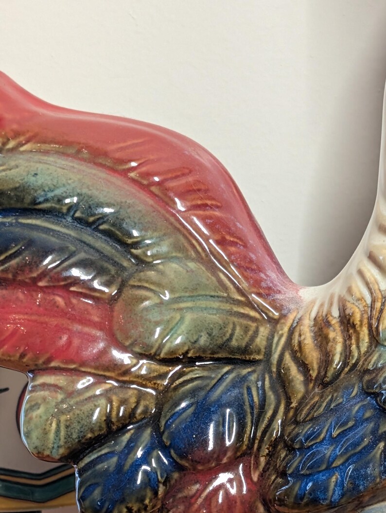 May include: Close-up of a ceramic rooster with a glossy finish. The rooster features a vibrant color palette, including red, blue, green, and brown. The details of the feathers are visible, adding texture to the design. The rooster is a decorative item.
