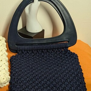 Puede incluir: Un bolso de mano de ganchillo azul marino con un asa oscura. El bolso tiene un patrón texturizado y un asa triangular. Un maniquí de mano blanco está en el fondo.