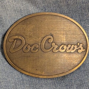 May include: Oval-shaped brass belt buckle with the words "Doc Crow's" in a cursive font. The buckle has a worn, vintage appearance, with visible scratches and a slightly tarnished finish. The background is a denim fabric.