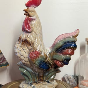 May include: A ceramic rooster figurine with a red comb and wattle, beige body, and colorful tail feathers. The rooster is standing on a base with green and blue accents. A decorative item for a kitchen or farmhouse decor.
