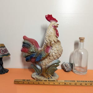 May include: A ceramic rooster figurine with a red comb and tail feathers in shades of blue, green, and red. The rooster is standing on a base with green accents. A wooden ruler with text is in front of the rooster.