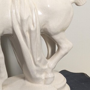 Puede incluir: Estatua de caballo de cer&aacute;mica color crema con un detalle de tela drapeada. El caballo est&aacute; posado con las patas en movimiento y la estatua tiene un acabado brillante. La escultura est&aacute; montada sobre una base rectangular.