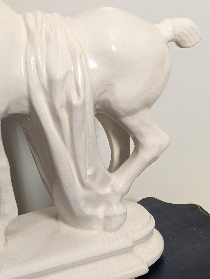 Puede incluir: Estatua de caballo de cer&aacute;mica color crema con un detalle de tela drapeada. El caballo est&aacute; posado con las patas en movimiento y la estatua tiene un acabado brillante. La escultura est&aacute; montada sobre una base rectangular.