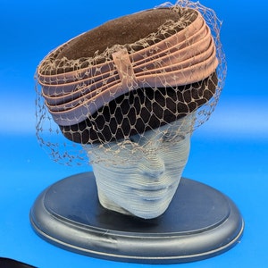 Puede incluir: Un sombrero de terciopelo marrón con un lazo de satén rosa y un velo de red negro. El sombrero está sobre un soporte negro.