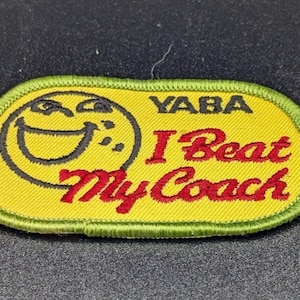 May include: A yellow embroidered patch with a green border. The patch features a smiling face and the text "YABA I Beat My Coach" in black and red lettering. The patch is rectangular with rounded corners.