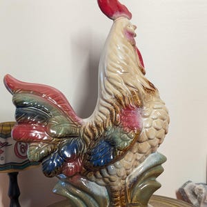 May include: A ceramic rooster figurine with a red comb and wattle, and a multi-colored tail. The rooster has a cream-colored body with brown and pink accents, and is perched on a base with green and brown details.