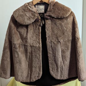 May include: A brown faux fur cape with a large collar and a  'King's' label. The cape has a  hood and a  front closure with two  decorative buttons.