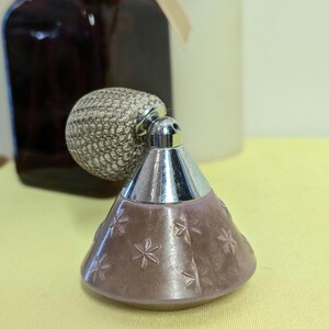 May include: A vintage perfume bottle with a silver-colored textured bulb atomizer and a cone-shaped body in a muted mauve color. The body is decorated with embossed star-shaped designs. The bottle has a silver-colored metal collar and nozzle.