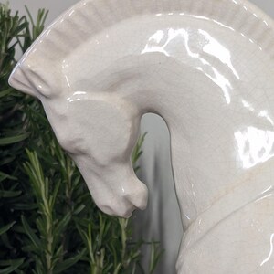 Puede incluir: Una escultura de cabeza de caballo de cer&aacute;mica color crema. La cabeza del caballo est&aacute; inclinada hacia abajo, con una melena y rasgos faciales detallados. La escultura tiene un acabado brillante y un efecto craquelado, lo que le da un aspecto vintage. Se ve follaje verde en el fondo.