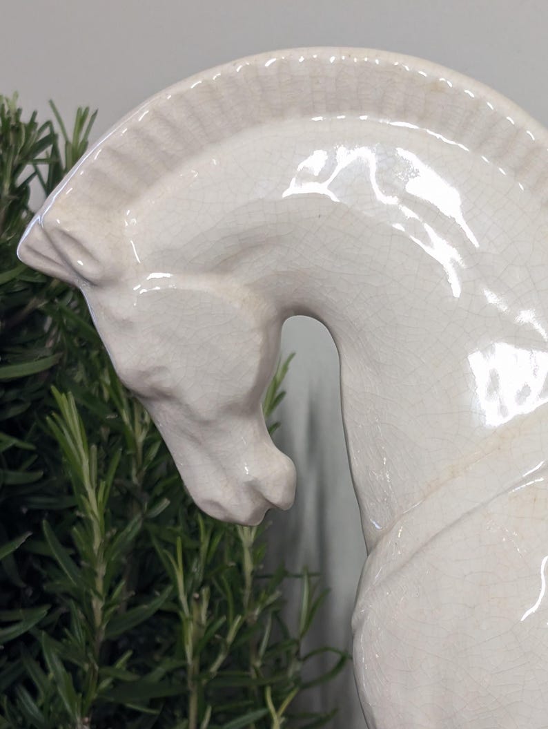 Puede incluir: Una escultura de cabeza de caballo de cer&aacute;mica color crema. La cabeza del caballo est&aacute; inclinada hacia abajo, con una melena y rasgos faciales detallados. La escultura tiene un acabado brillante y un efecto craquelado, lo que le da un aspecto vintage. Se ve follaje verde en el fondo.