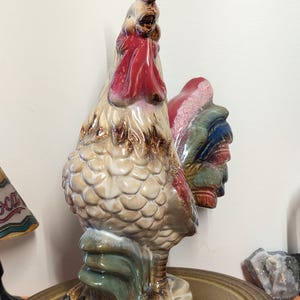 May include: A ceramic rooster figurine with a red comb and wattle, tan body, and colorful tail feathers. The rooster is standing on a base with green and blue accents. The figurine is a decorative item.