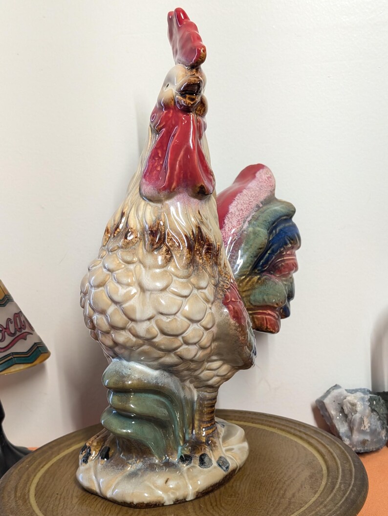 May include: A ceramic rooster figurine with a red comb and wattle, tan body, and colorful tail feathers. The rooster is standing on a base with green and blue accents. The figurine is a decorative item.