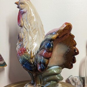 May include: A ceramic rooster figurine with a red comb and wattle, cream-colored body, and colorful tail feathers. The rooster is perched on a round, wooden base. The figurine has a glossy finish and is likely vintage.