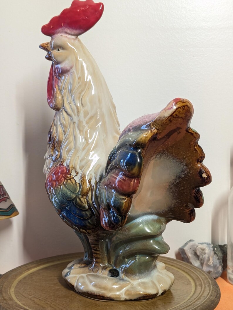 May include: A ceramic rooster figurine with a red comb and wattle, cream-colored body, and colorful tail feathers. The rooster is perched on a round, wooden base. The figurine has a glossy finish and is likely vintage.