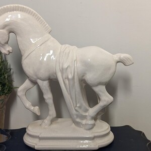 Puede incluir: Una estatua de caballo de cer&aacute;mica de color crema. El caballo est&aacute; representado en una pose din&aacute;mica, con una pata levantada y cubierto con tela. La estatua est&aacute; montada sobre una base decorativa. El dise&ntilde;o general es elegante y cl&aacute;sico.
