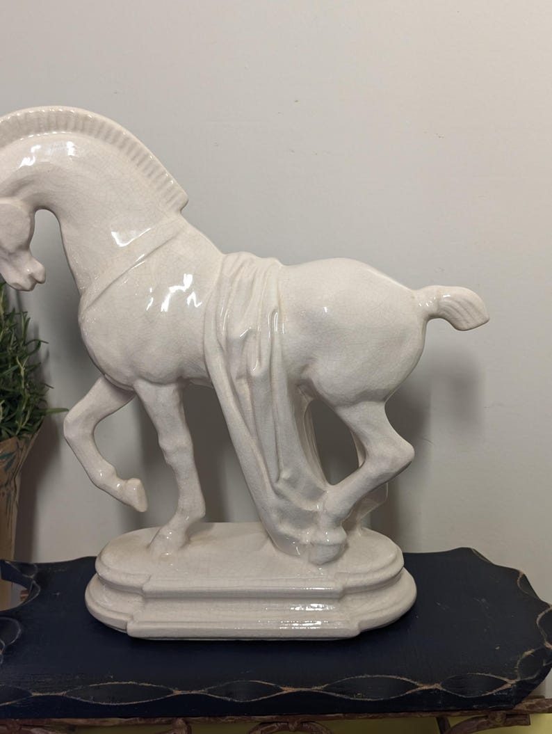 Puede incluir: Una estatua de caballo de cer&aacute;mica de color crema. El caballo est&aacute; representado en una pose din&aacute;mica, con una pata levantada y cubierto con tela. La estatua est&aacute; montada sobre una base decorativa. El dise&ntilde;o general es elegante y cl&aacute;sico.