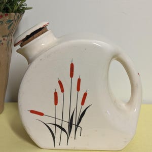 May include: Cream-colored ceramic pitcher with a handle and a spout, featuring a design of red cattails and black leaves. The pitcher has a cork stopper and is likely vintage or antique.