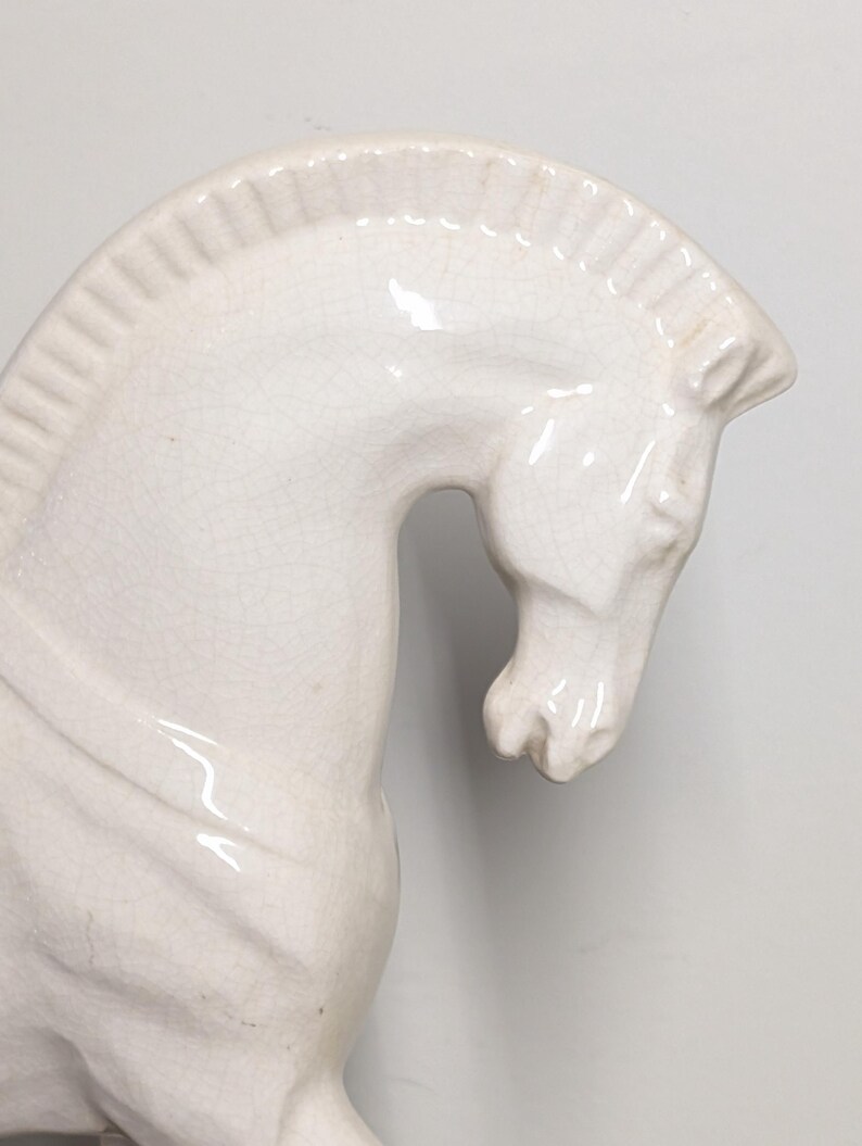 Puede incluir: Figura de caballo de cer&aacute;mica color crema. El caballo se representa de perfil, con la cabeza y el cuello arqueados. La figura tiene un acabado brillante y detalles de craquelado visibles, agregando textura. Un art&iacute;culo decorativo.